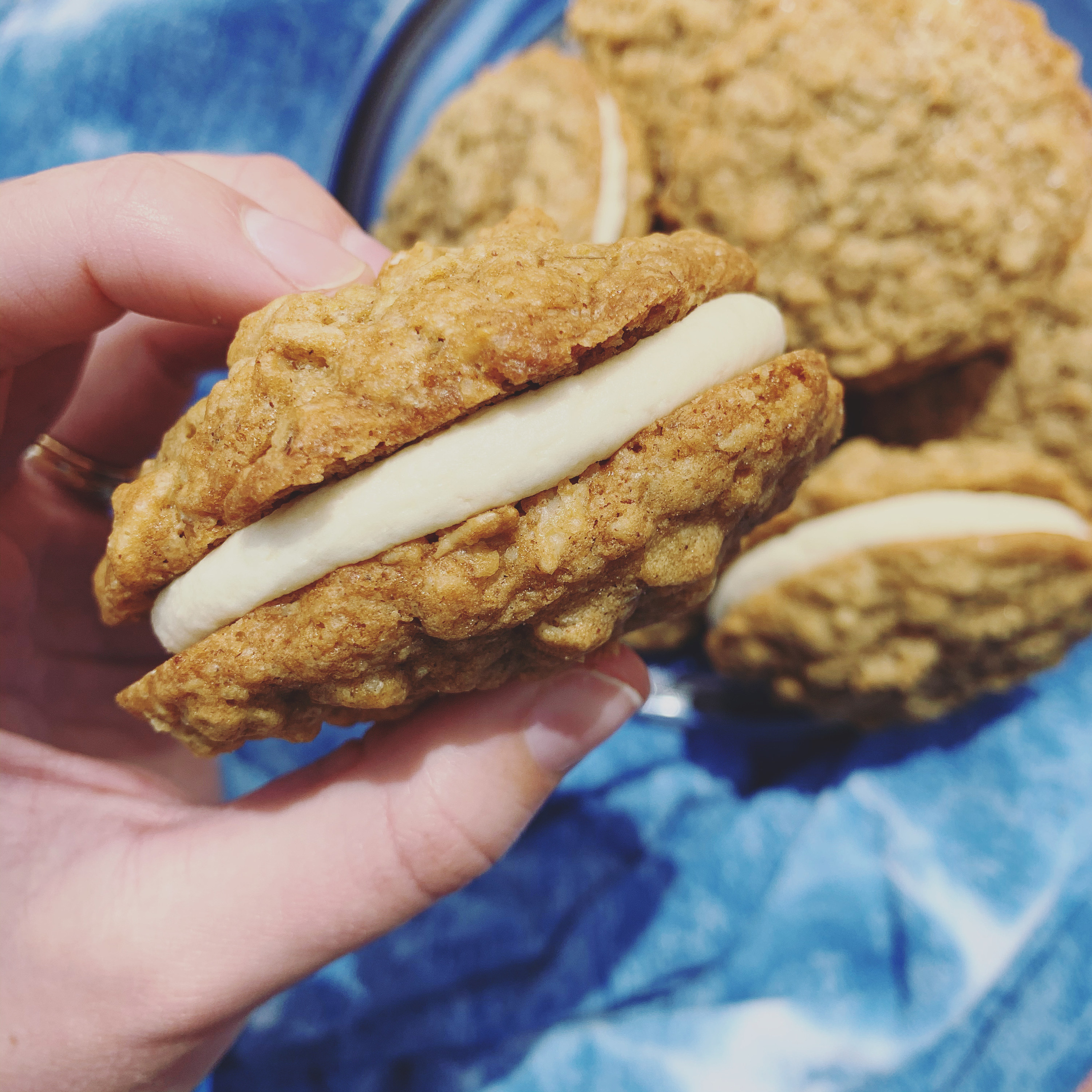 A hand holding a two golden oatmeal biscuits sandwiched together with buttercream icing