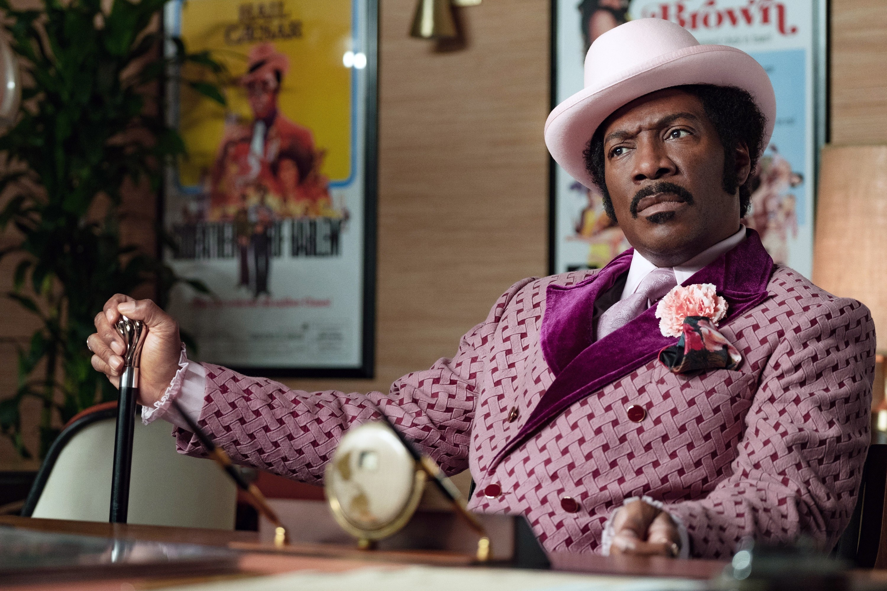 still image of actor eddie murphy in the film dolemite, he is sitting down with a cane in one hand wearing a burgundy suit and light pink hat