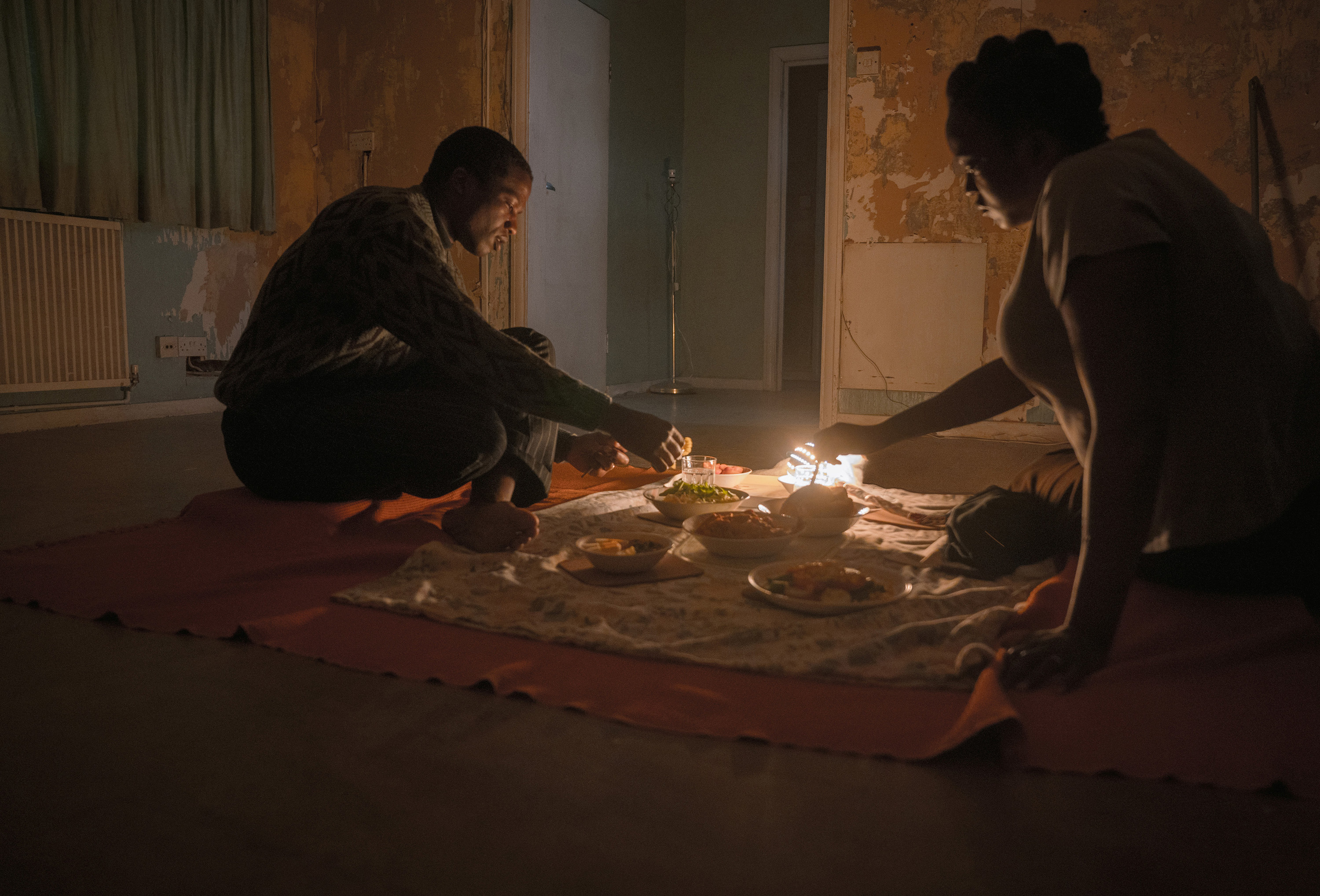 still image from the movie his house, a couple are sitting on the floor getting ready to eat in their new apartment 