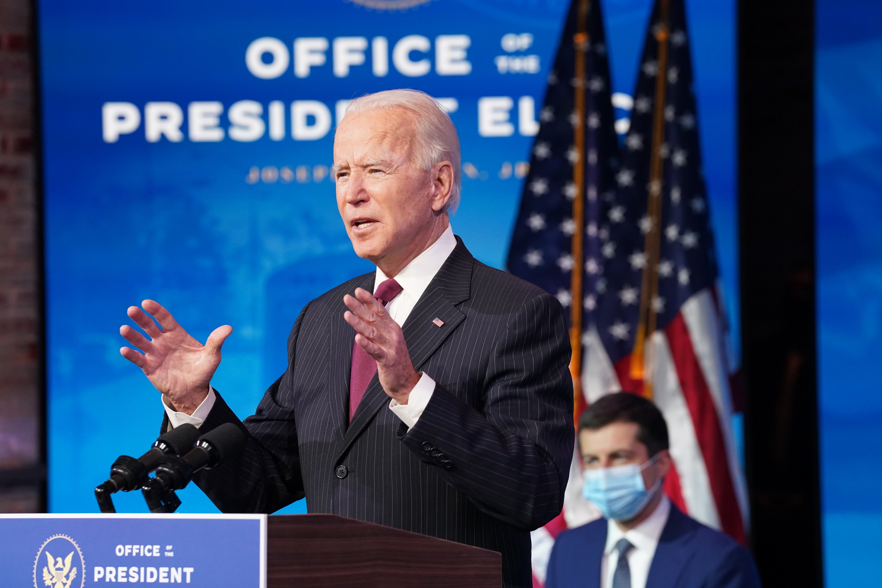 Joe Biden speaks at a podium as Pete Buttigieg sits behind him wearing a face mask