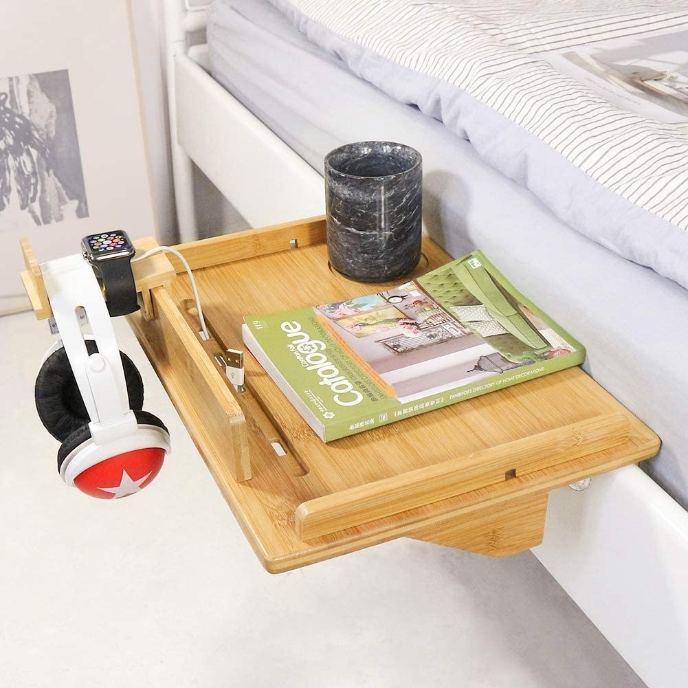 bedside caddy on the side of a bed with a glass of water, a book, and a headphone set next to an apple watch