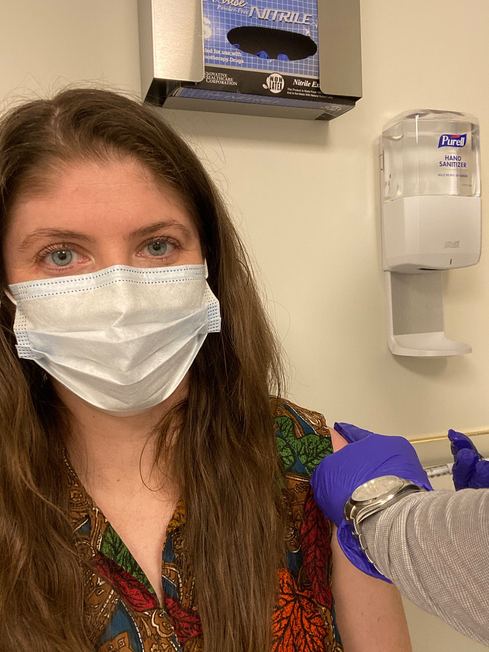 The writer, sitting and wearing a paper face mask, gets an injection in her left arm