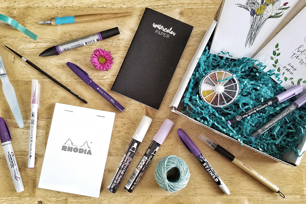 An open box with an array of notebooks, markers, pens, and other lettering tools 