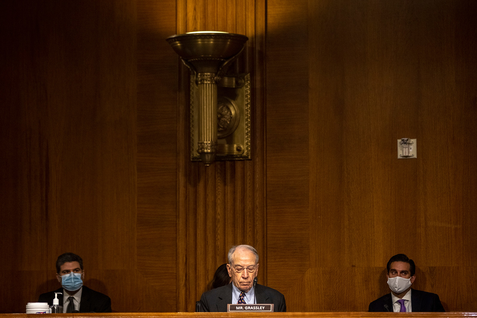 Three men sit behind a long bench with Senator Chuck Grassley at the center sitting behind a placard that reads &quot;Mr Grassley&quot;