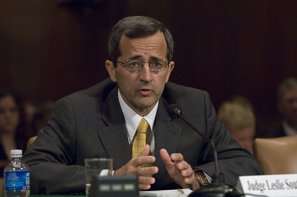 A man sits behind a table and speaks into a microphone