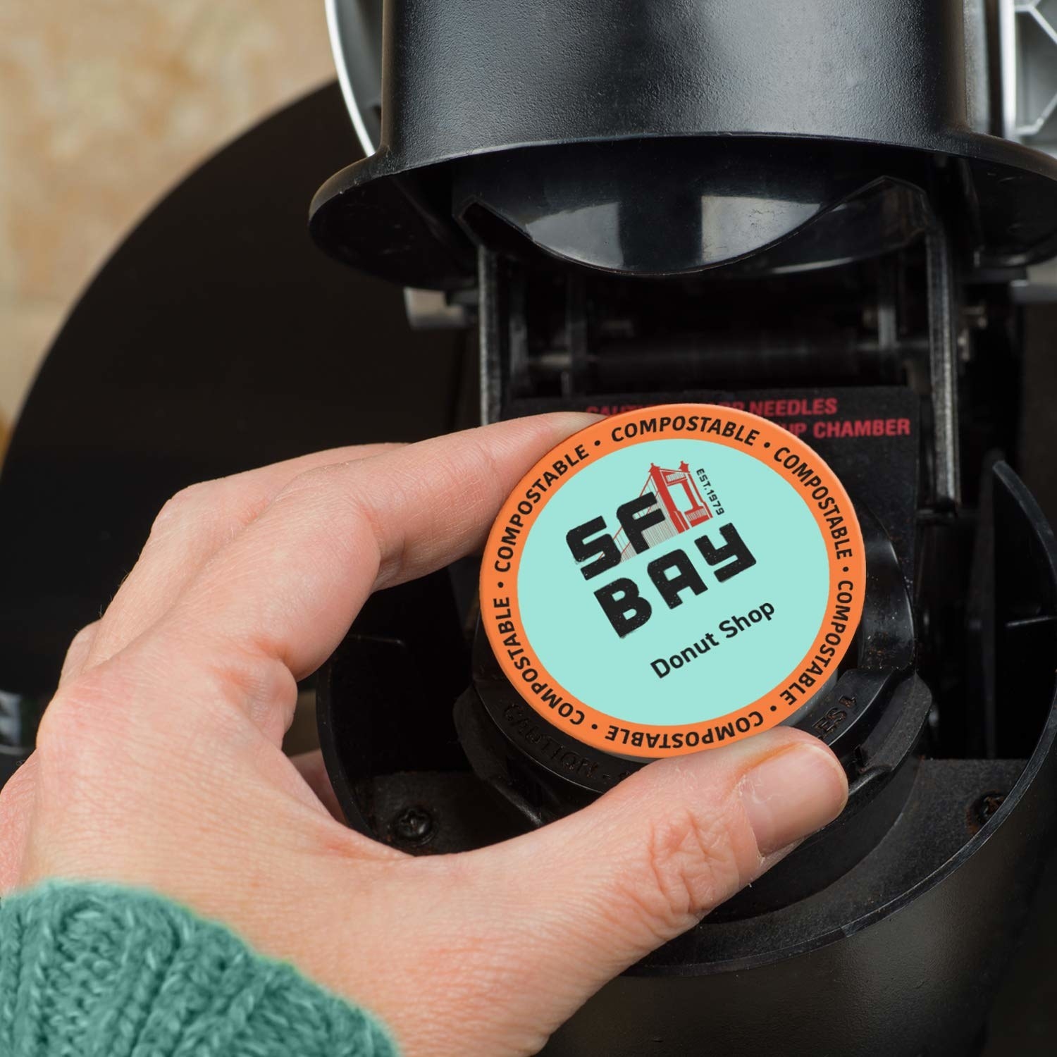 Hand holds compostable coffee pod that says &quot;SF Bay Donut Shop&quot; on the front and puts it inside Keurig-like coffee machine