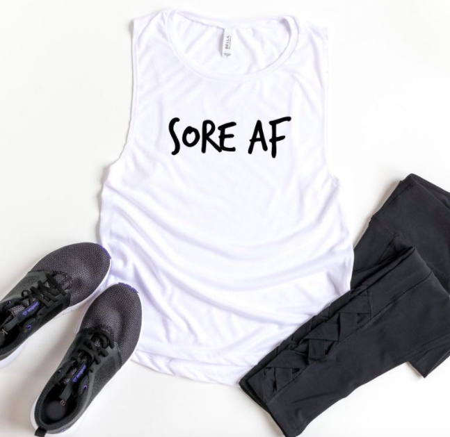 A flatlay of the muscle tank that reads sore af next to a pair of running shoes and leggings