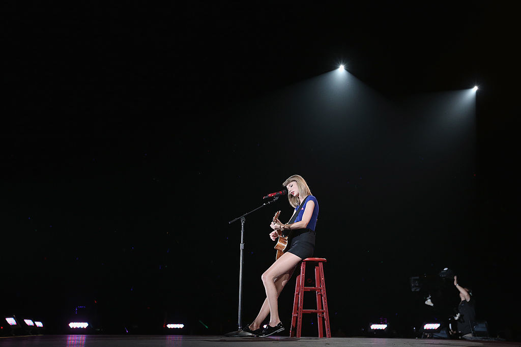 a woman with bangs and shoulder-length hair sits on a wooden stool on a large stage. she sings into a microphone while playing her guitar, looking down at it