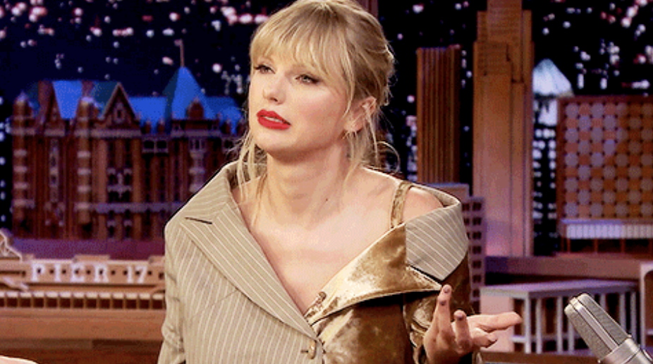 a woman with bangs and a messy ponytail sits in a chair on a late night talk show. she is shrugging, mouth lopsided, teeth slightly showing, eyes almost closed