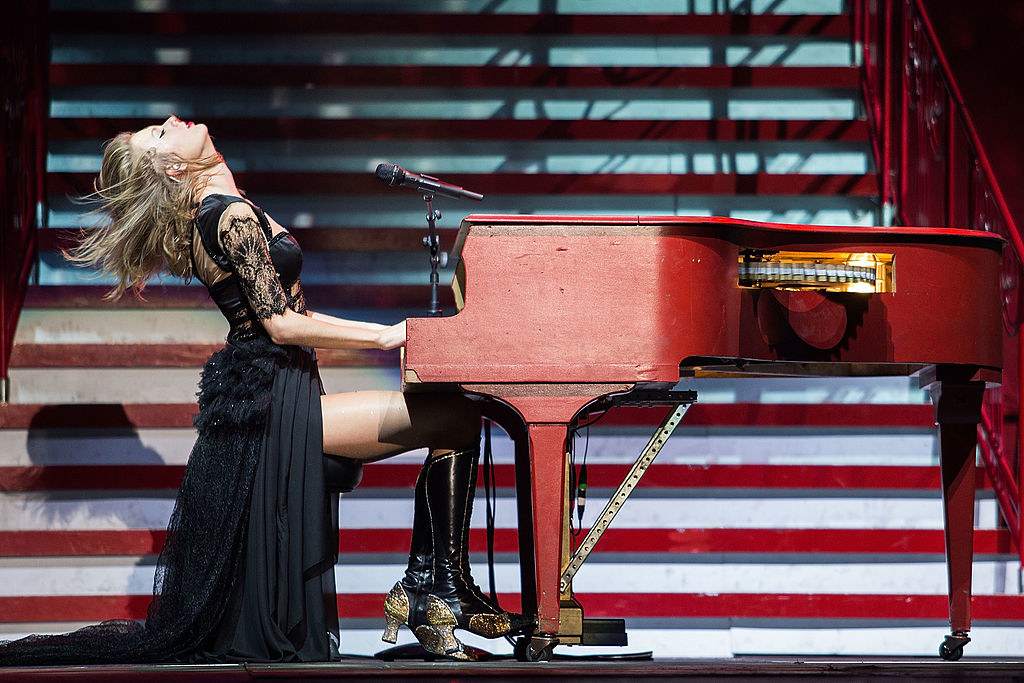a woman in a long dress with lacy sleeves sits upon a large stage. she is throwing her head back, hair flowing, hands on a piano. 