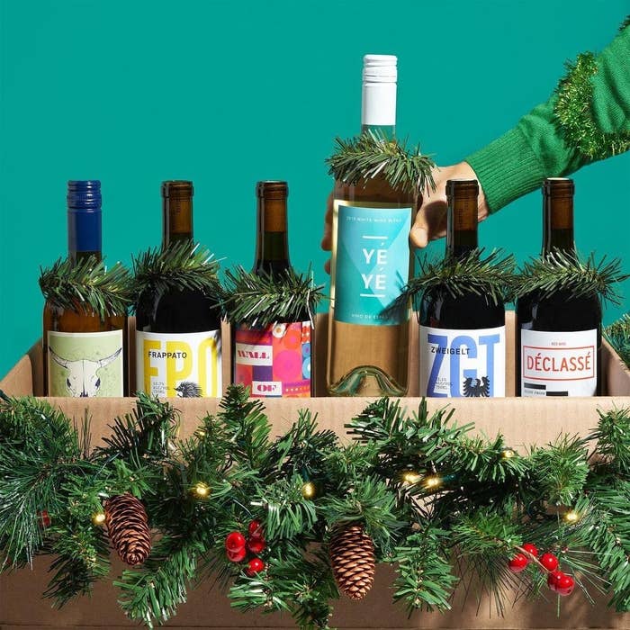 A hand putting a bottle of wine into a box filled with over bottles of wine decorated with tinsel and pine cones 