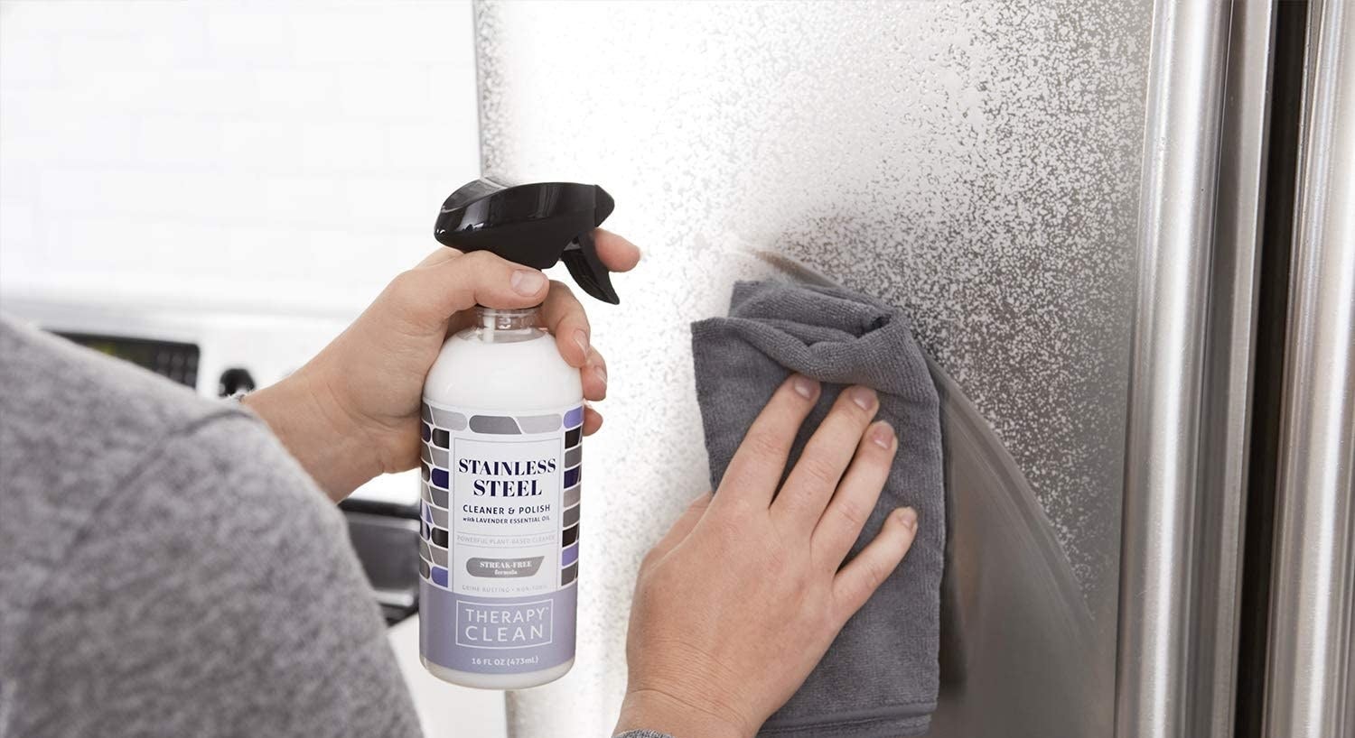 Hands spraying the white cleaner on a fridge, then wiping with a microfiber cloth