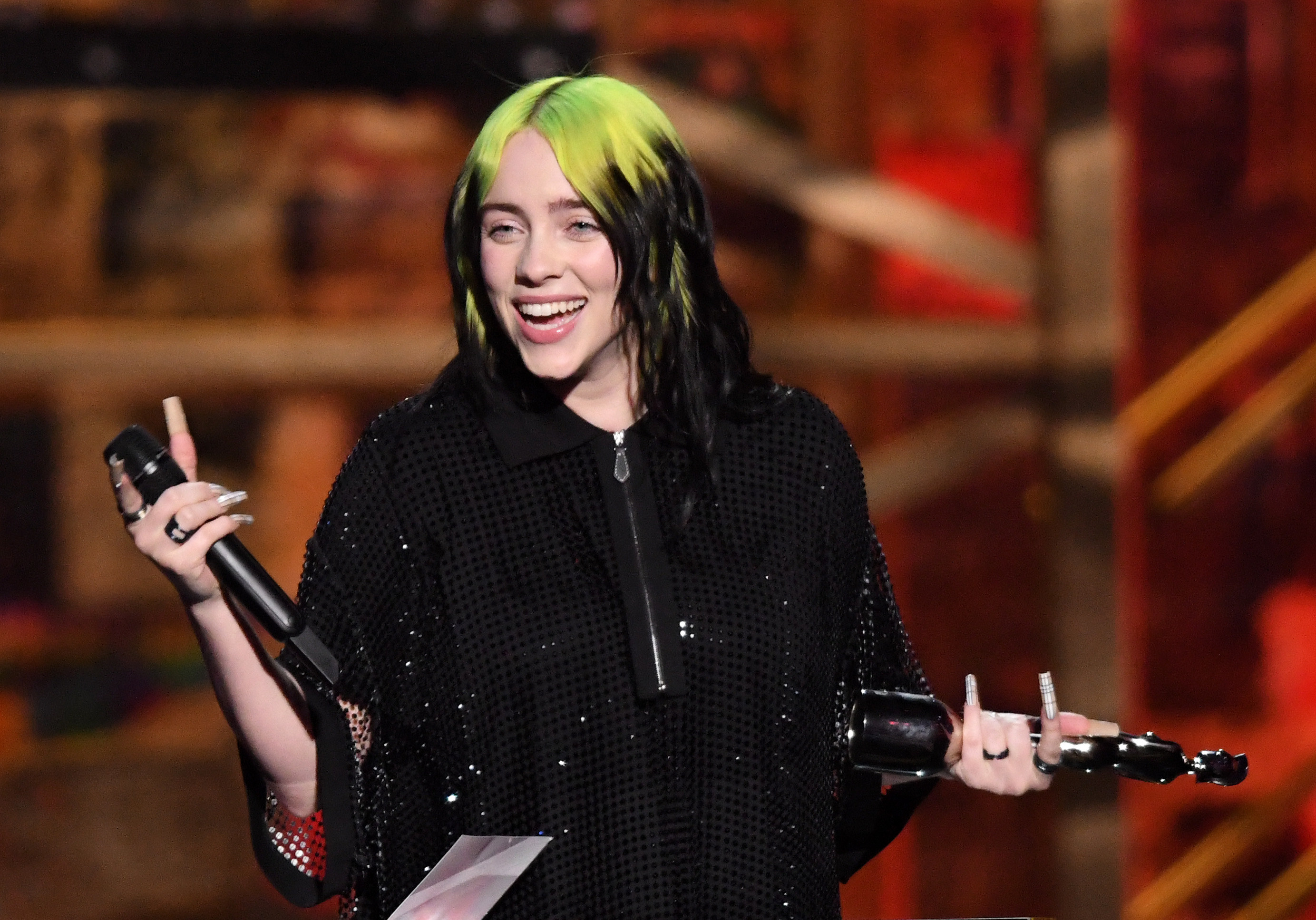 Billie Eilish smiling, holding a microphone and an award