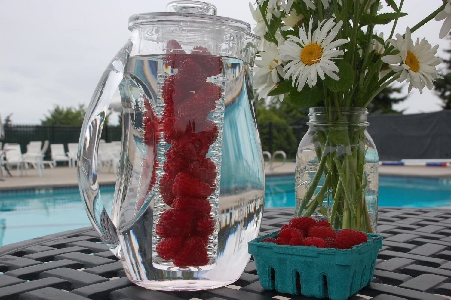 A fruit infusing pitcher on a patio table next to a basket of raspberries and a vase of daisies 
