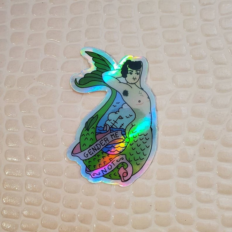 a holographic sticker that reads &quot;gender me not&quot; and shows a merperson with top surgery scars 