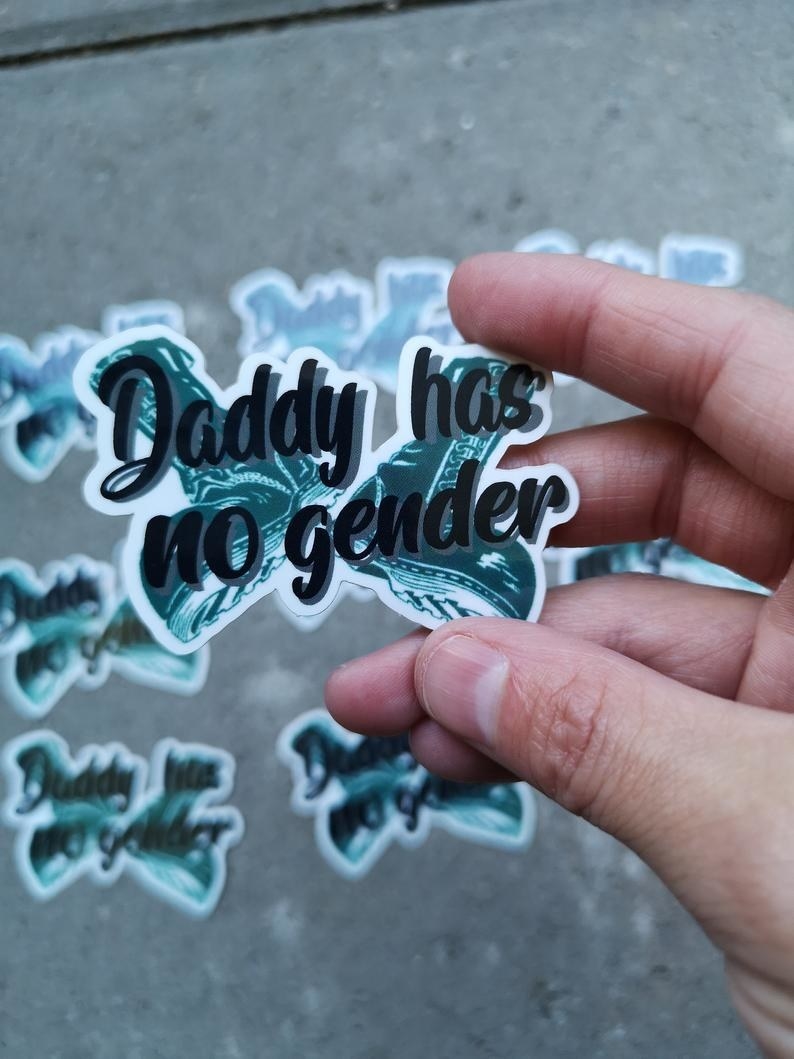 a hand holding the &quot;Daddy has no gender&quot; sticker with the wording over a pair of black boots