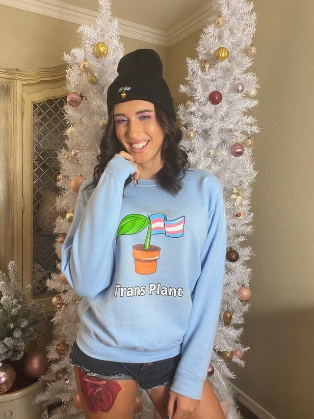Rose modeling the trans plant sweater in blue which has a little sprout growing out of a pot with a trans flag on the right side of the plant 