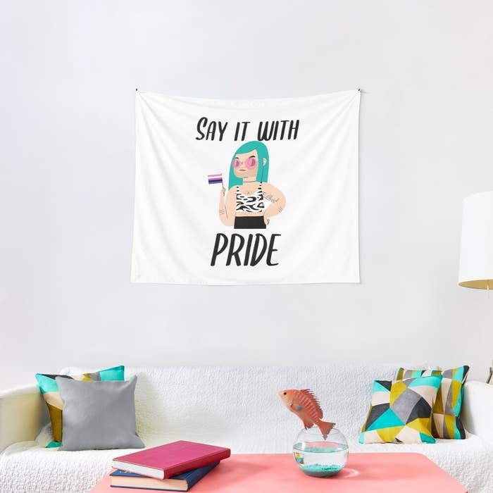 the tapestry which says &quot;Say it with Pride&quot; hanging on a wall with an illustration of a gender diverse person holding the bisexual pride flag