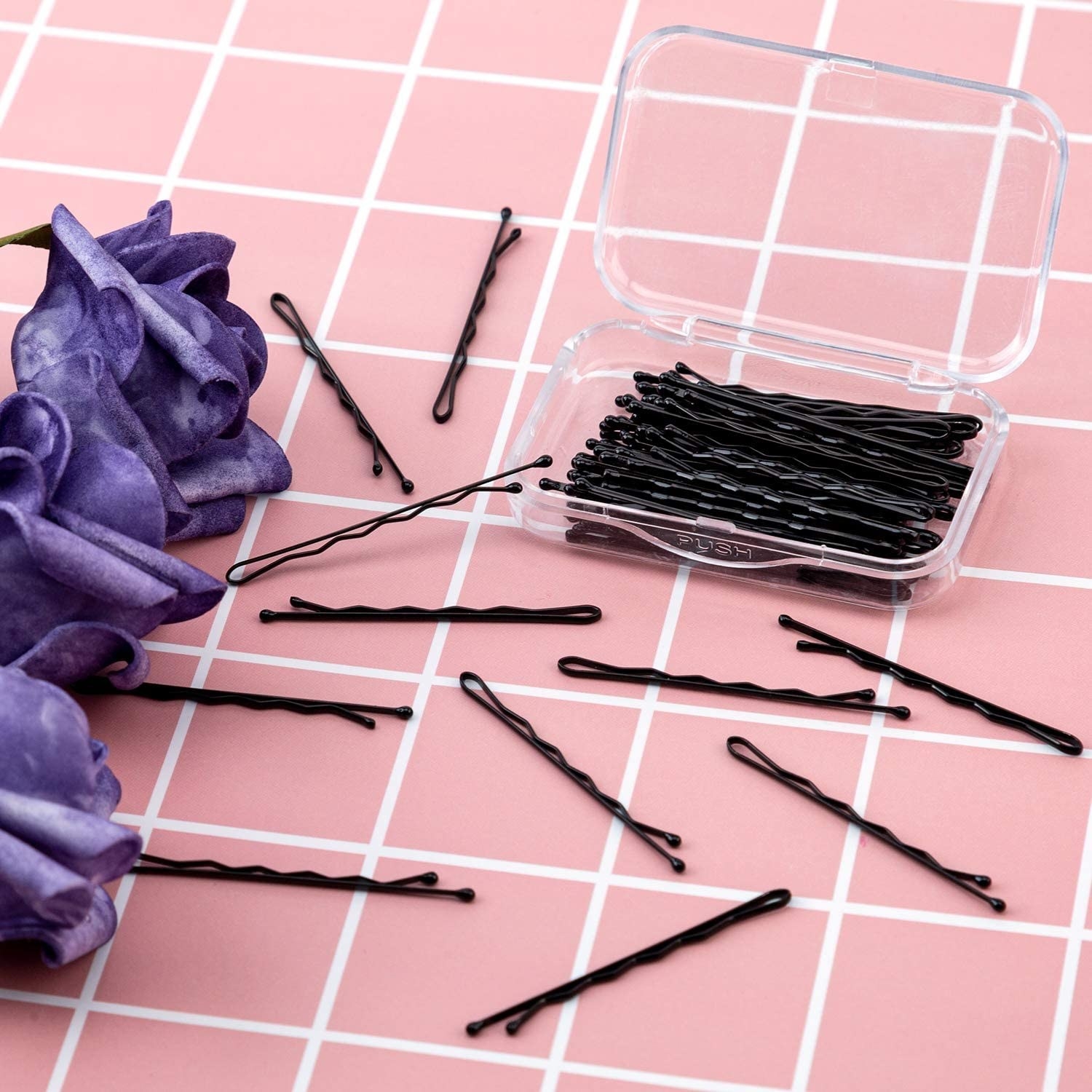 The open case of bobby pins surrounded by several bobby pins and flowers