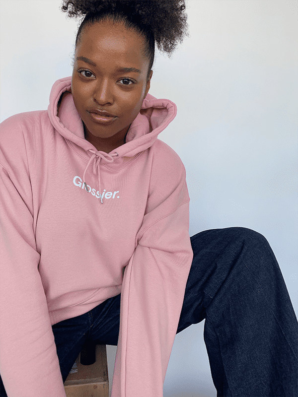 Model wears pink Glossier hoodie with white Glossier logo on the front