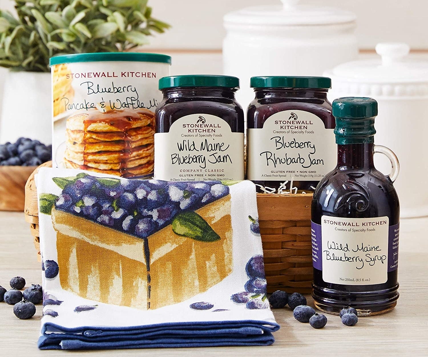 A collection of jams, pancake mix, and maple syrup in a basket with a tea towel 