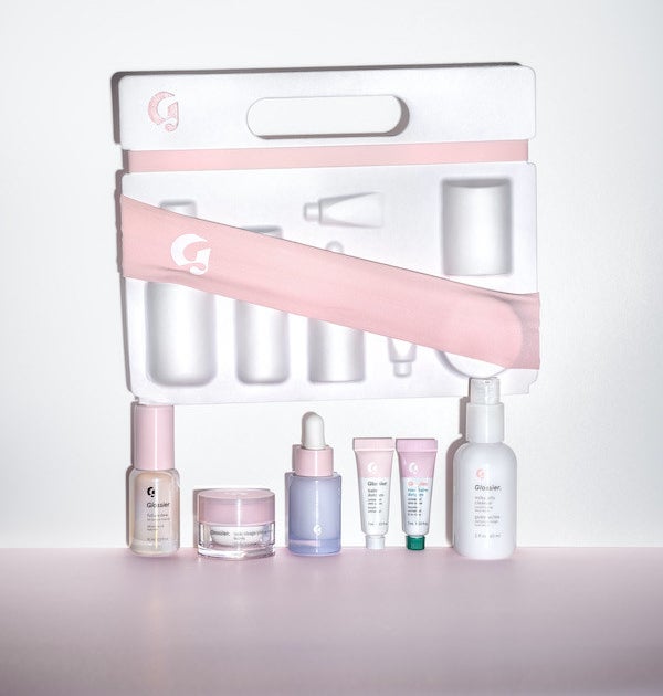 Mini clear and pink Glossier skincare products arranged in a neat line below a white box and pink headband