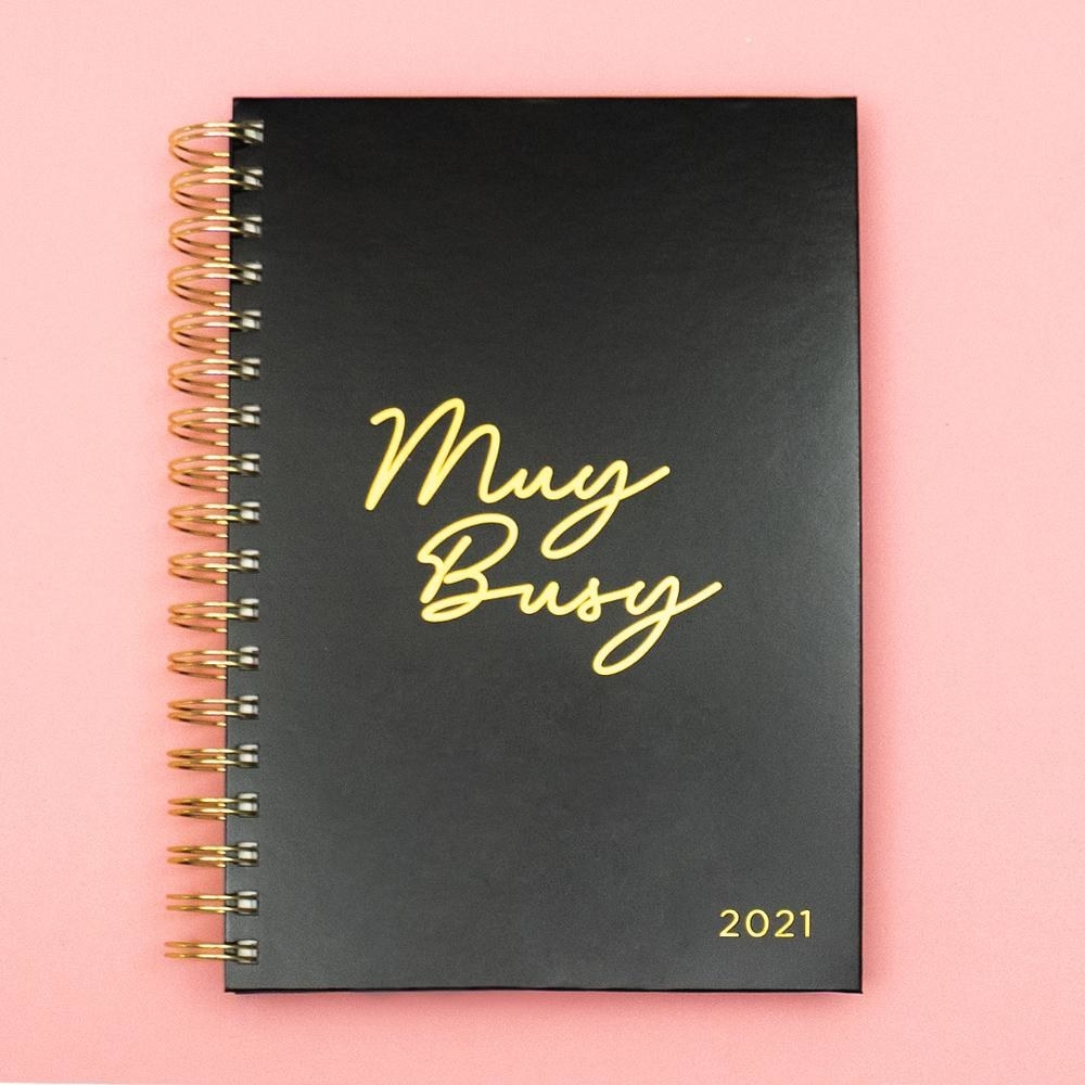 a black planner with a gold spiral spine that says "Muy Busy" on gold on the front