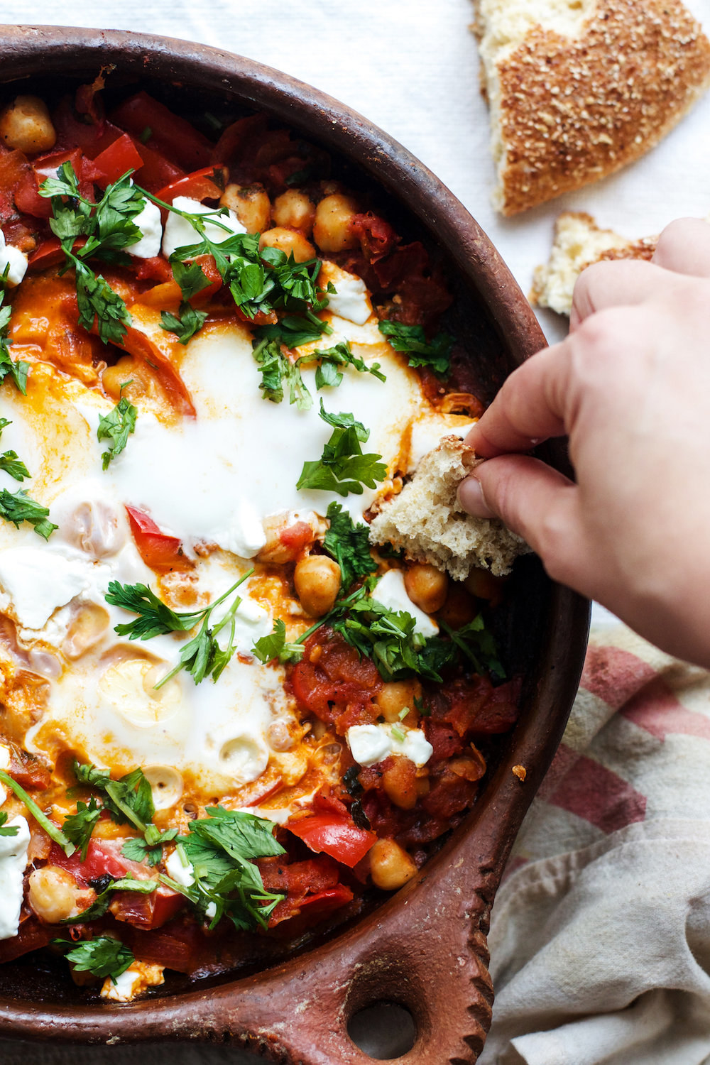 Someone dunking bread into a skillet of shakshuka with chickpeas, goat cheese, and herbs.