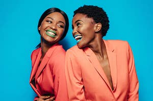 two women in matching burnt orange suits wearing green and blue lipstick while looking at each other smiling and laughing