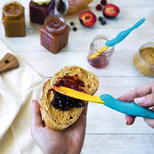 Hand uses platapus-shaped jar scraper spatula to spread jam on piece of bread with peanut butter