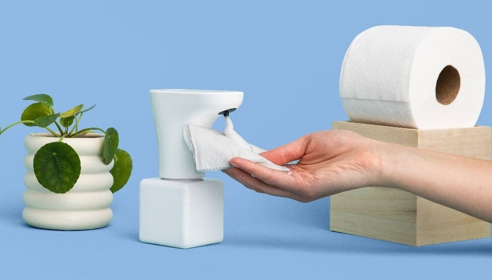A hand holding some toilet paper with foam on it under the Fohm dispenser next to a roll of toilet paper and a potted plant