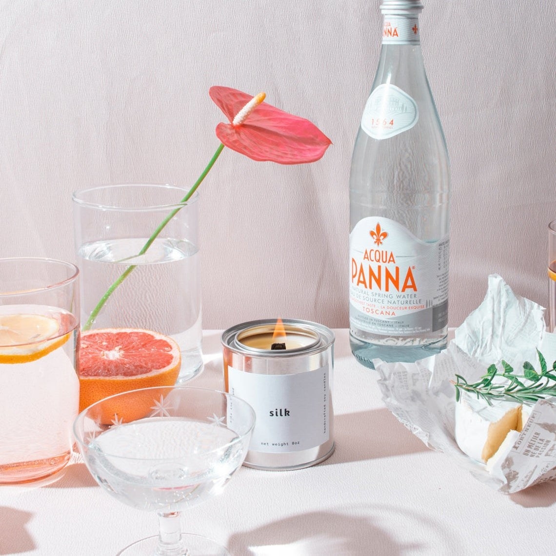 A lit candle in a metal tin with cups and of sparkling water beside it