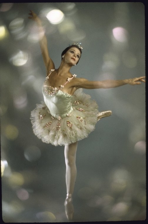 A ballerina in costume stands on one leg with glittering lights