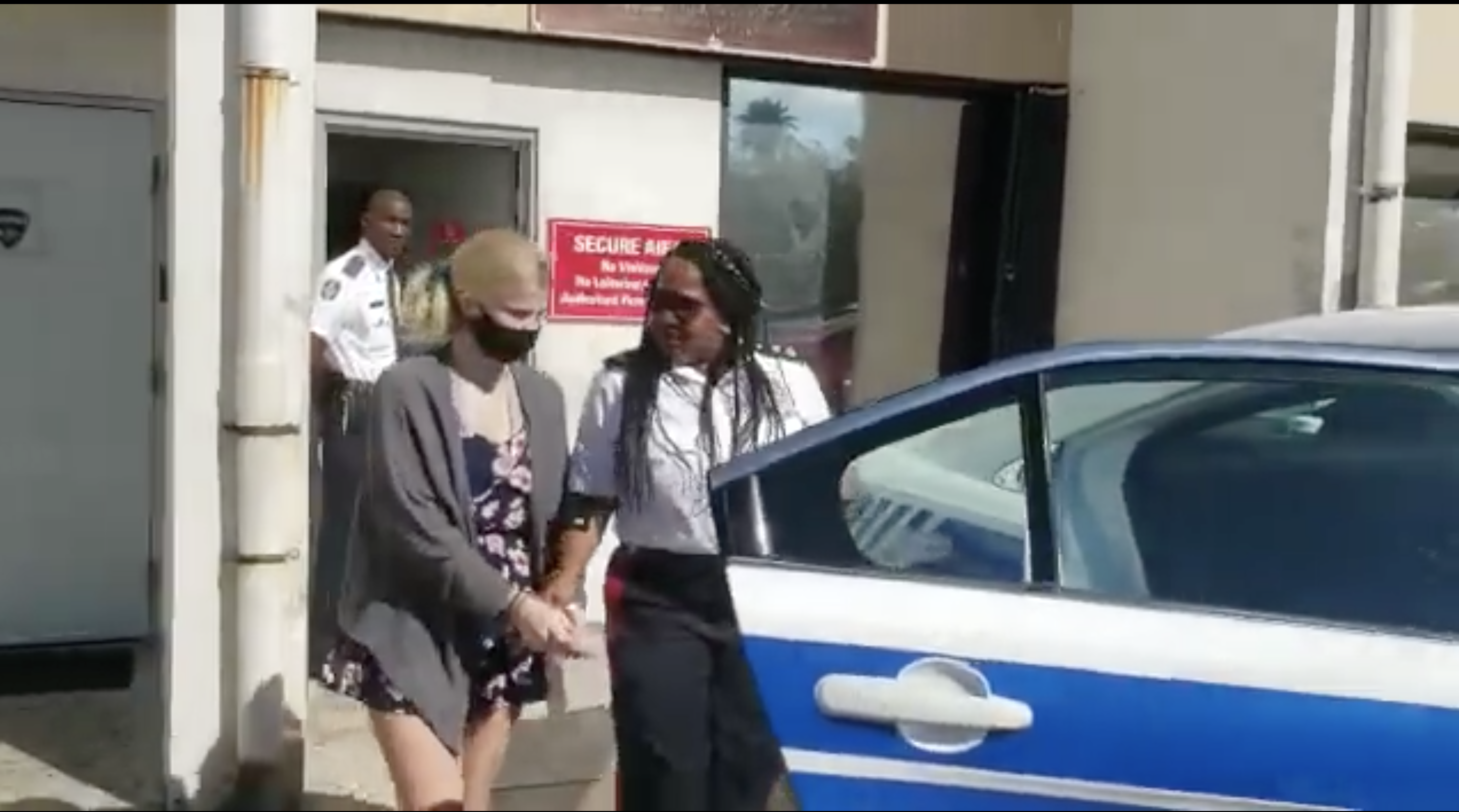 A police officer walks with Skylar Mack, handcuffed and wearing a face mask, to a police car