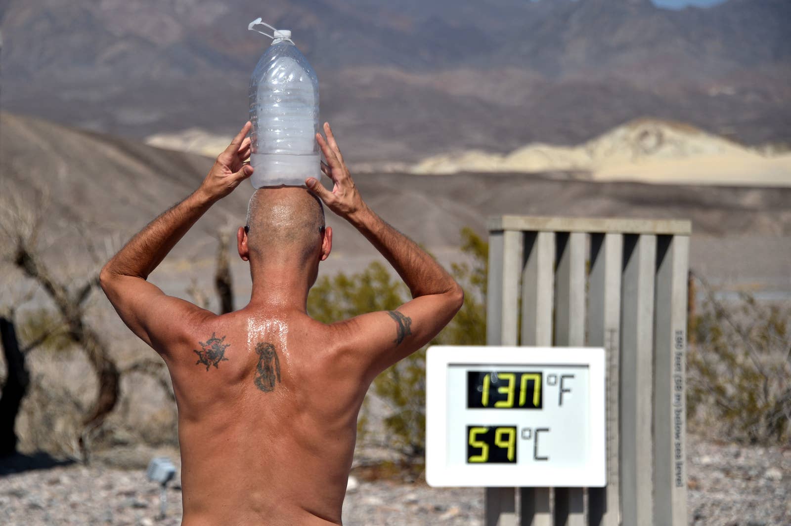 A shirtless man sweats with a frozen gallon of water on his head, standing in front of a thermometer that reads "130 degrees Fahrenheit, 59 degrees celsius"