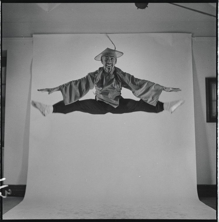 A man wearing a conical hat and sporting a long, fake mustache and goatee leaps in the air in front of a backdrop for a studio photo shoot