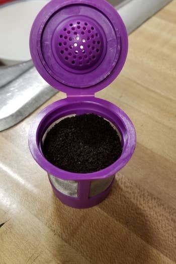 A purple K-cup with a hinged lid and stainless steel mesh sides filled with ground coffee 