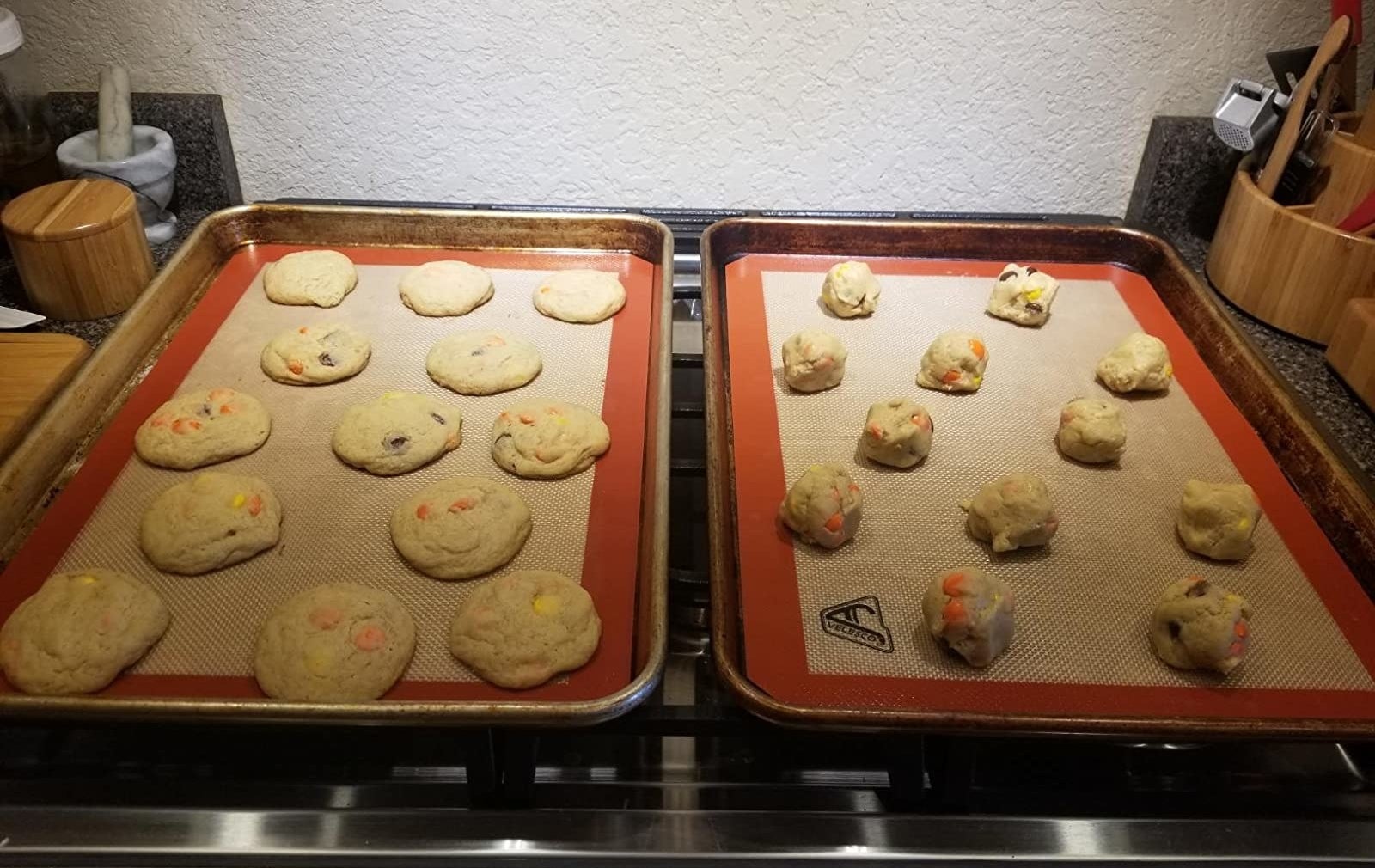 A reviewer photo of two baking sheets lined with the silicone mats and topped with baked cookies on one and balls of uncooked cookie dough on the other 
