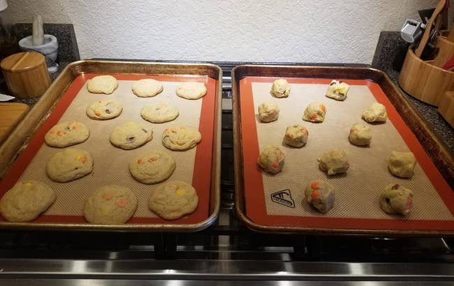 A reviewer photo of two baking sheets lined with the silicone mats and topped with baked cookies on one and balls of uncooked cookie dough on the other 