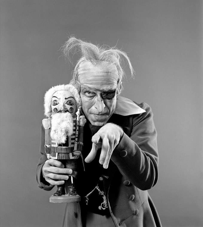 A man in old-age makeup wears an eye patch, thinning tufts of hair, holds a Nutcracker toy, and points at the camera