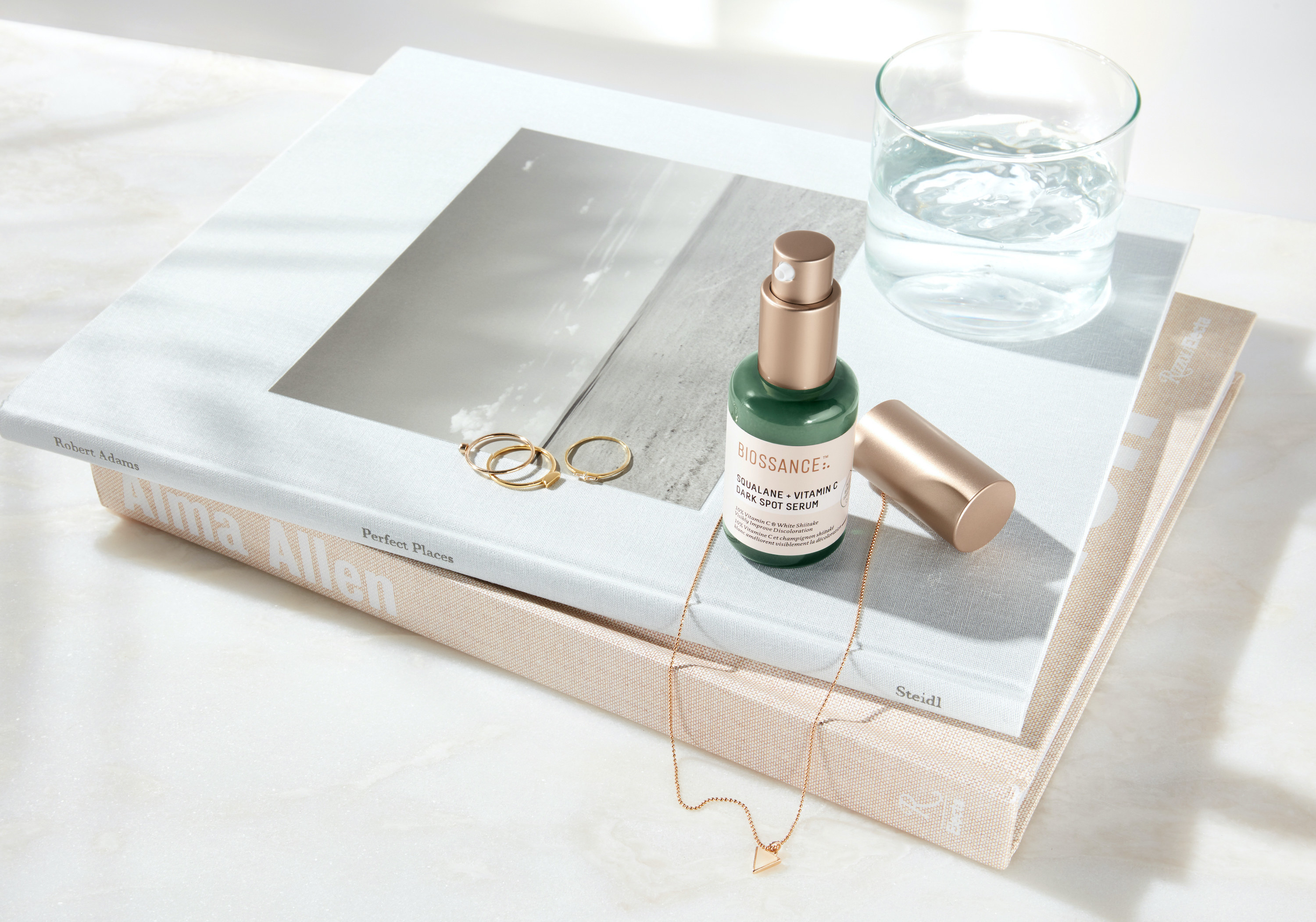 The Squalane and Vitamin C Dark Spot Serum in green bottle with a metallic pump top sitting on a stack of books next to a gold necklace and three rings 