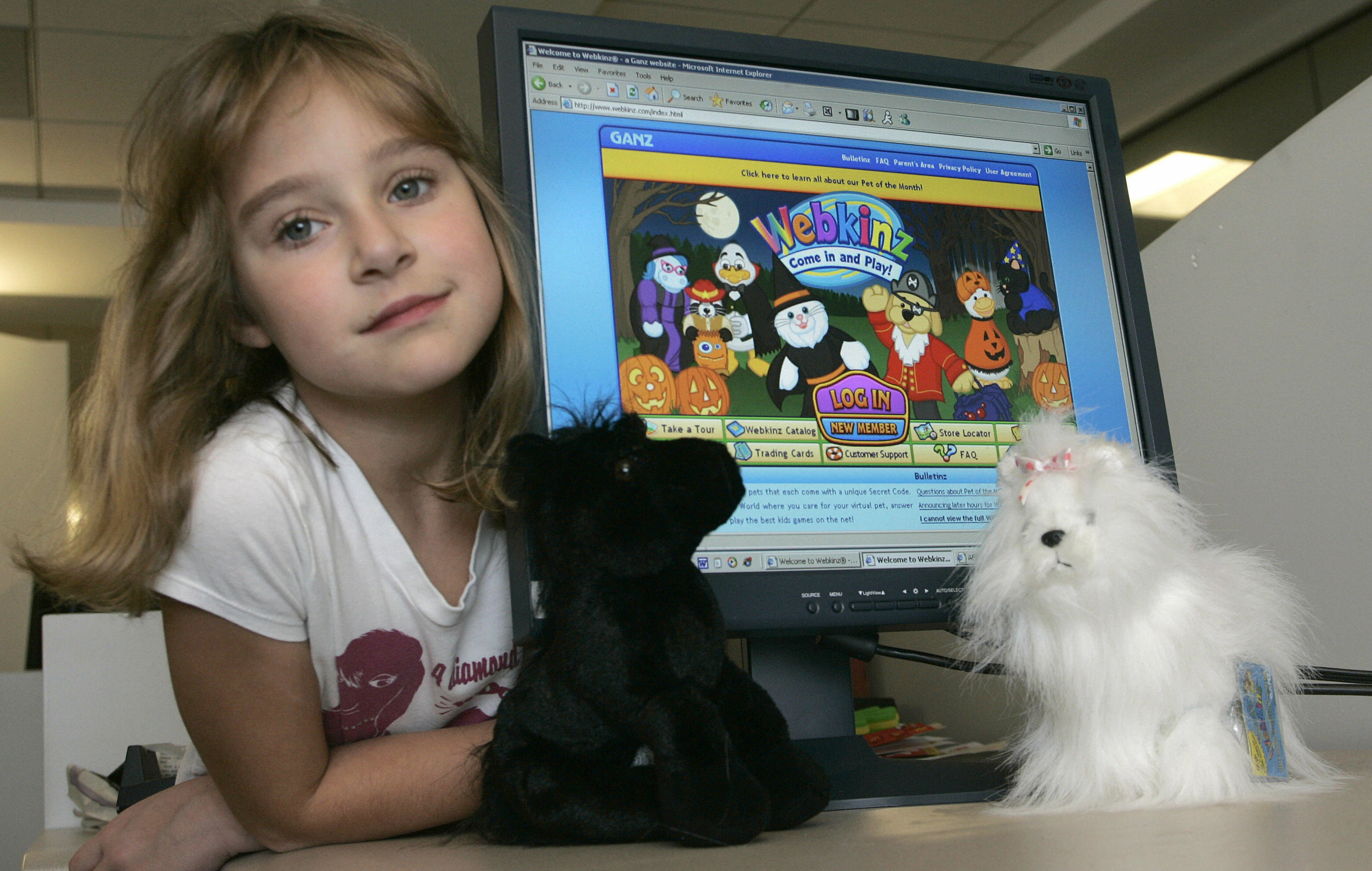 A young girl poses next to a computer showing the Webkinz website and two stuffed Webkinz 