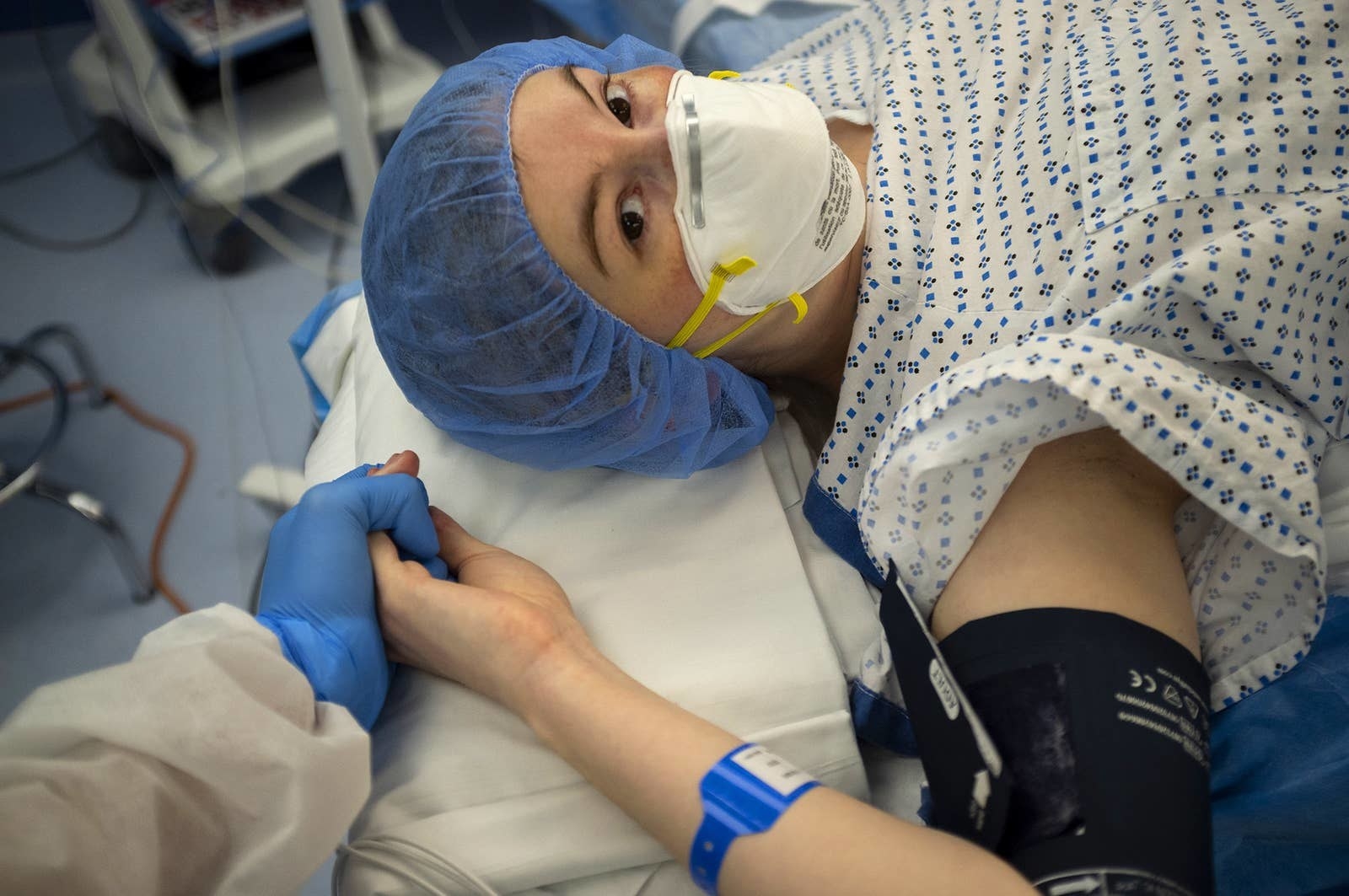 A woman in a mask and a hairnet on a hospital bed holds someone&#x27;s hand and looks at the camera