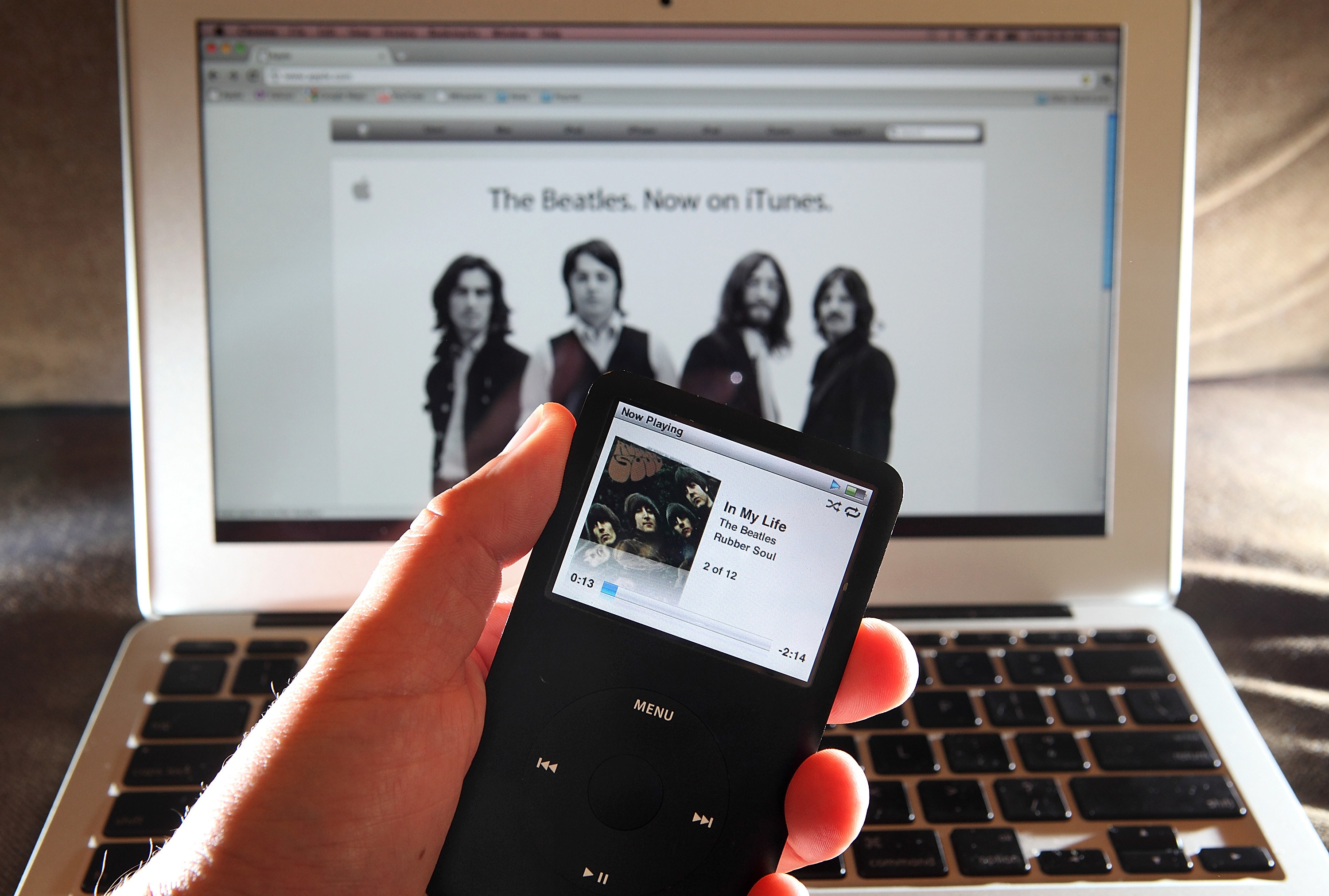 A person holds an iPod playing the Beatles in front of a Mac with iTunes open