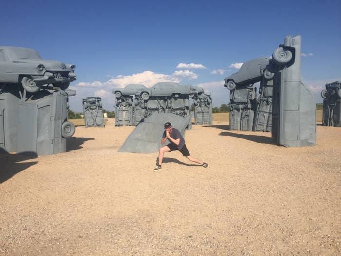 a man poses in front of sculptures made from old cars painted grey.