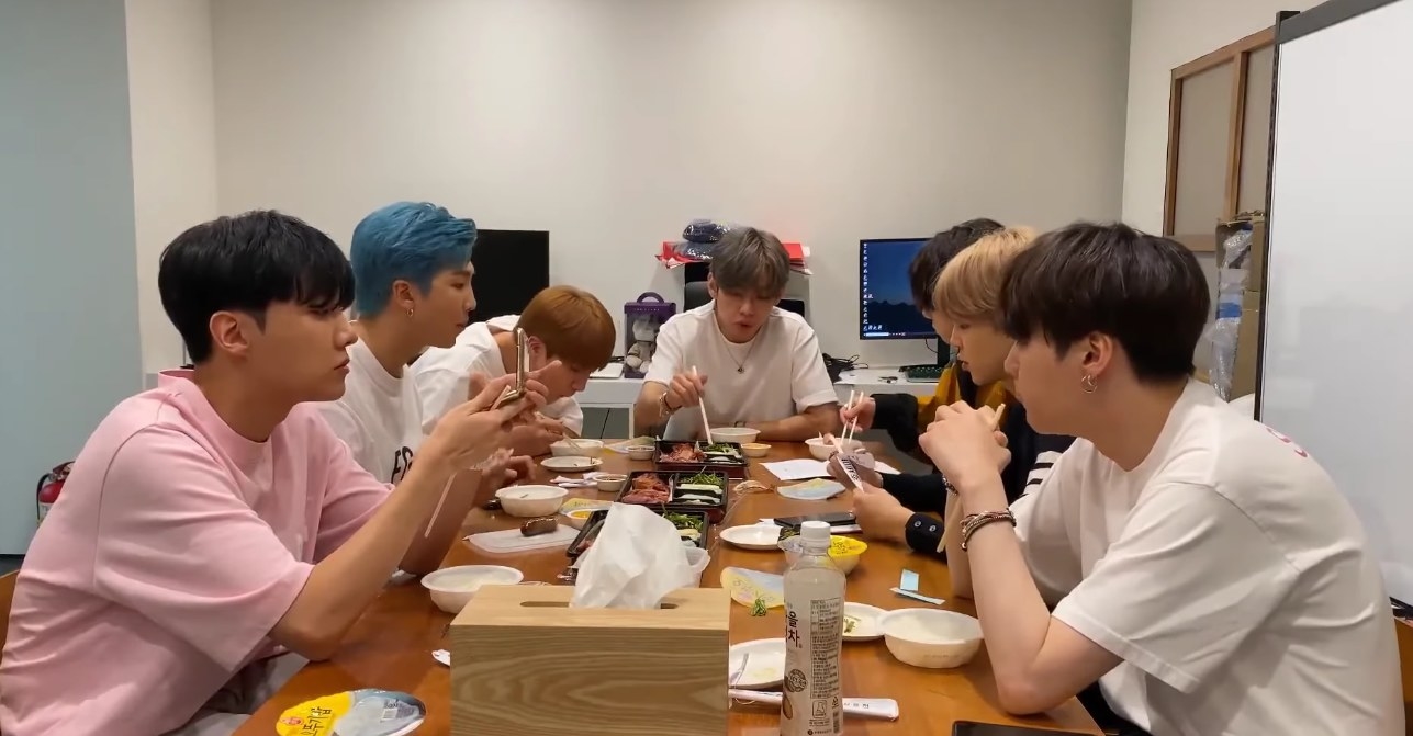 The BTS members sit around a meeting table eating and talking