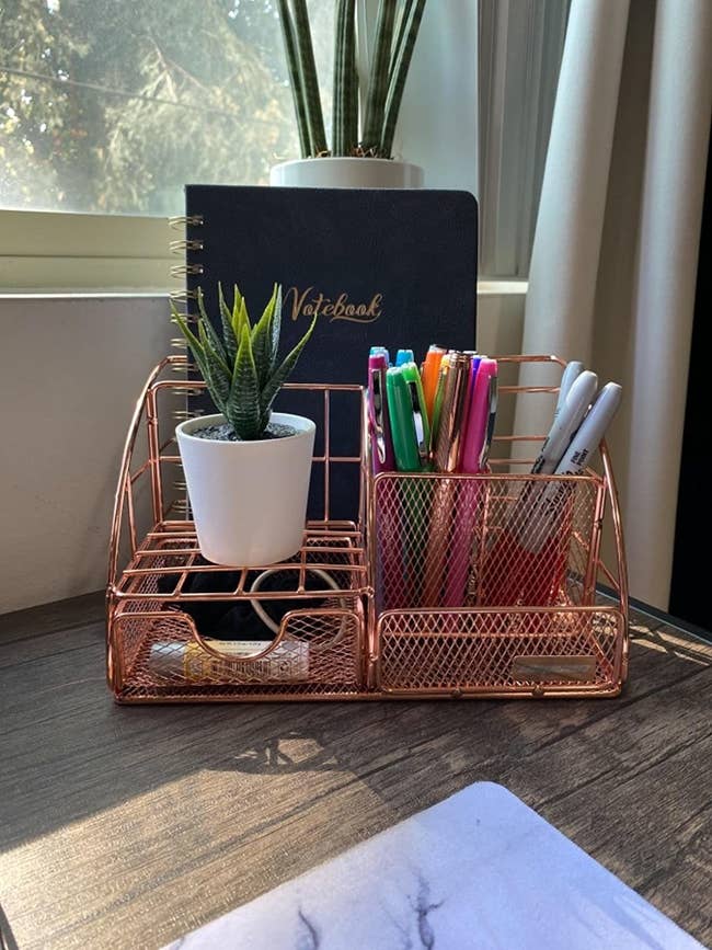 reviewer image of the rose gold desk organizer in the corner of a customer's desk holding a small succulent, pens, and a notebook