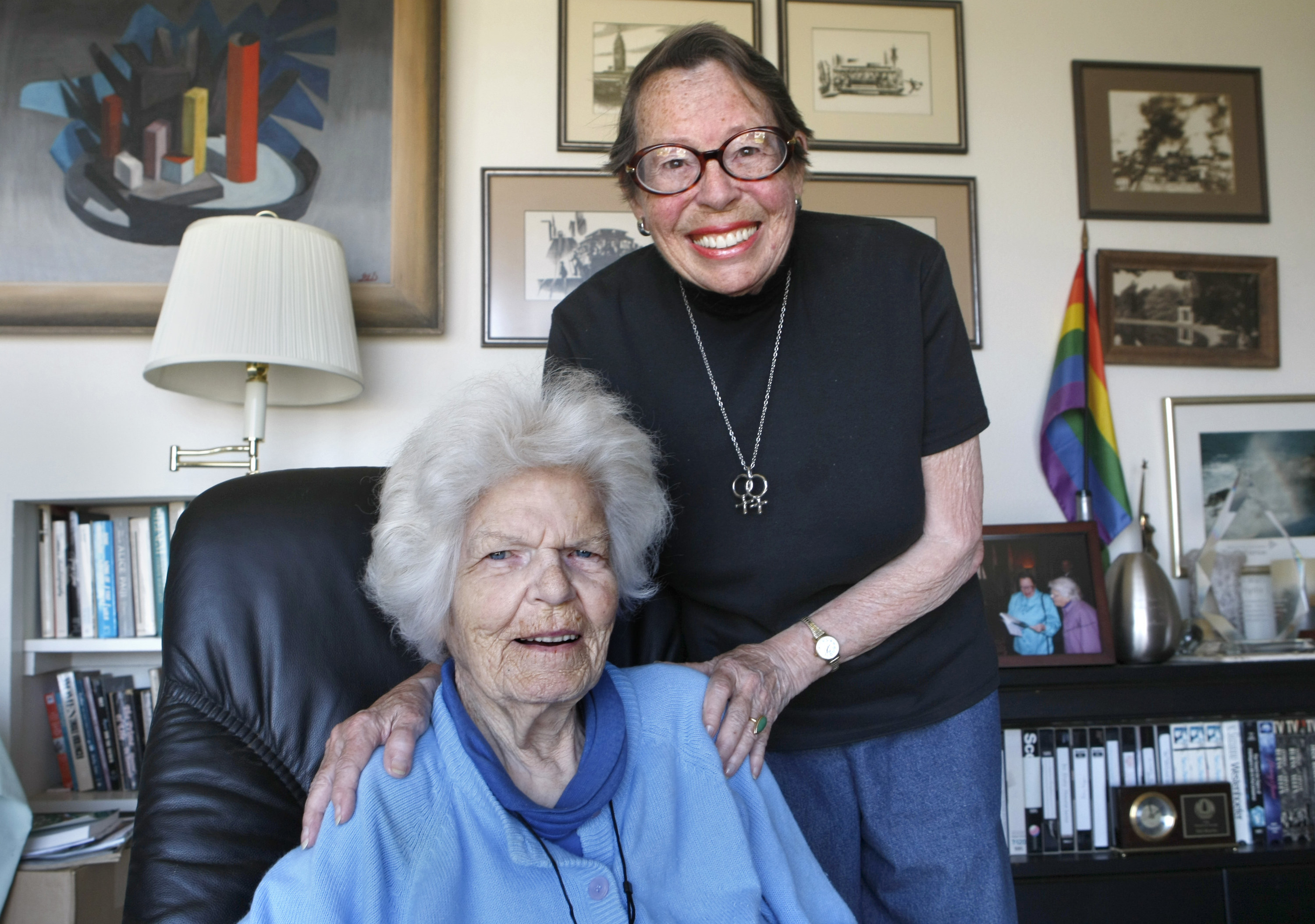 Two older women in their living room with a pride flag in the background