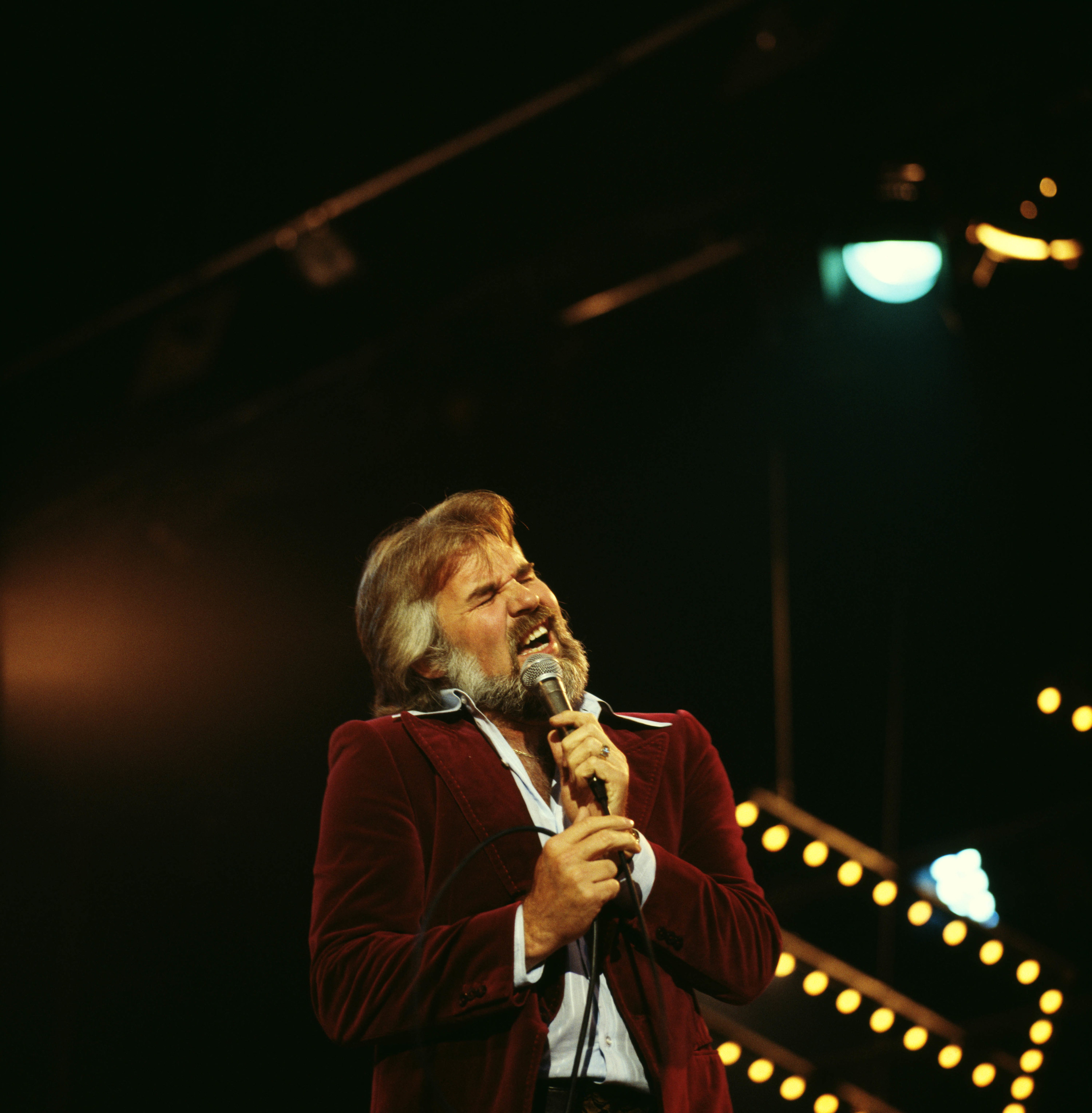 Rogers in a velvet suit singing into a microphone on stage with lights behind him