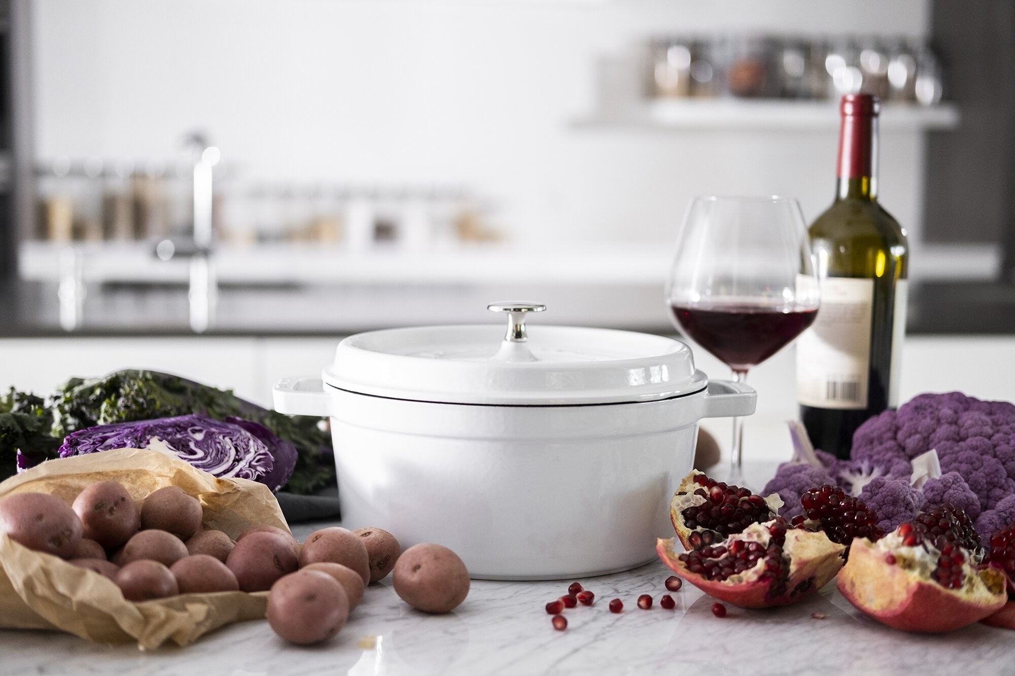 A white cast iron pot surrounded by ingredients and a glass of wine on a kitchen countertop 